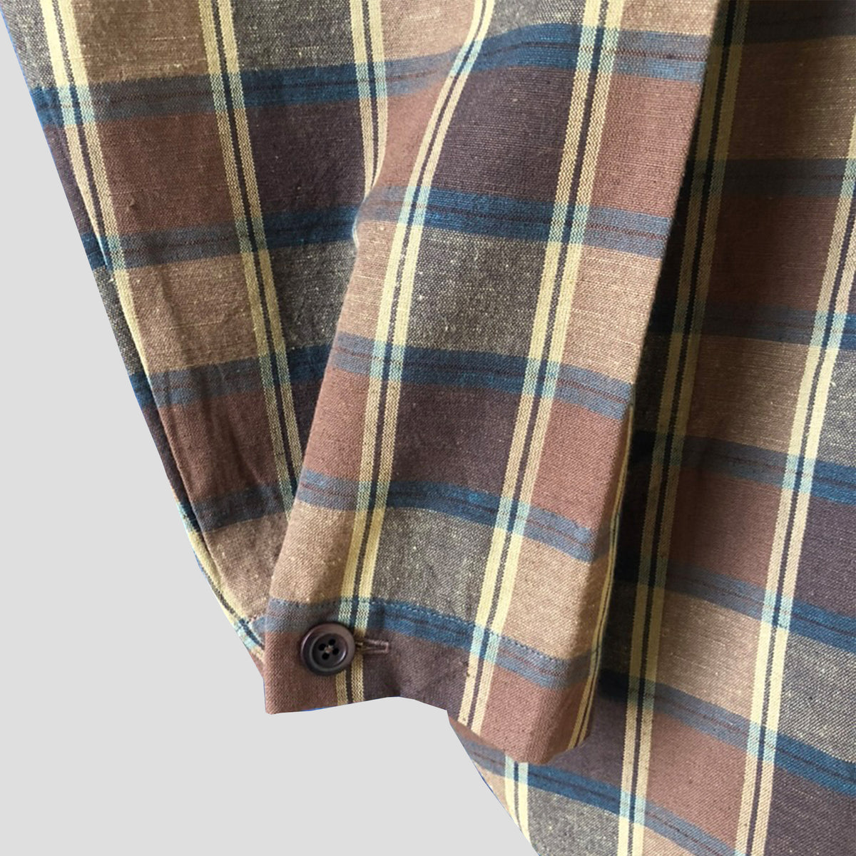 A close-up of a plaid shirt or jacket sleeve showing the fabric texture, button closure, and pattern details in brown, beige, and blue.