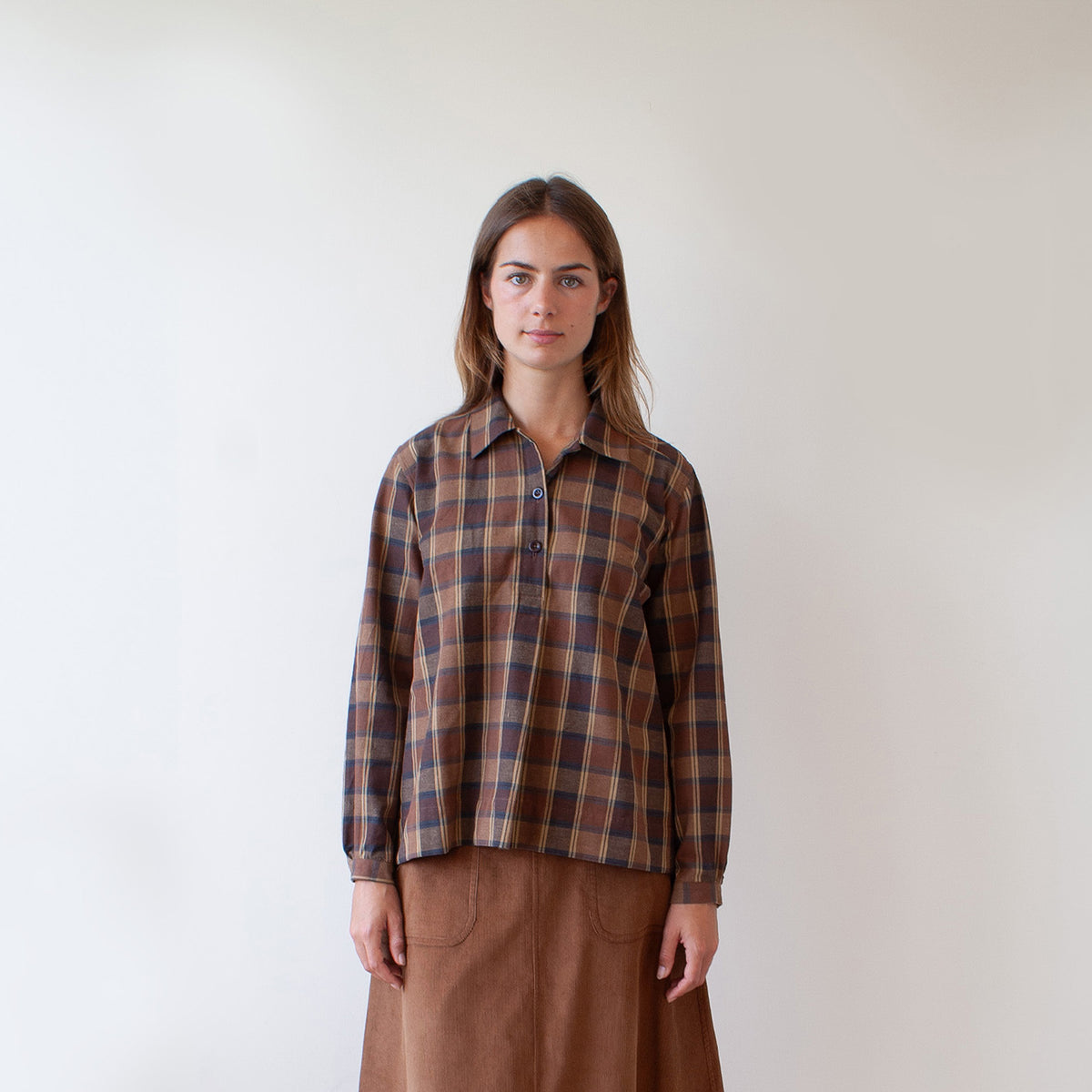 A woman stands against a plain off-white wall wearing a brown and beige plaid button-up shirt with a collar and a brown skirt.