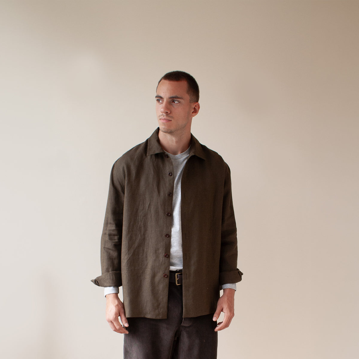 A man wearing a dark brown linen button-up shirt jacket over a white t-shirt and dark trousers, standing against a plain beige background.