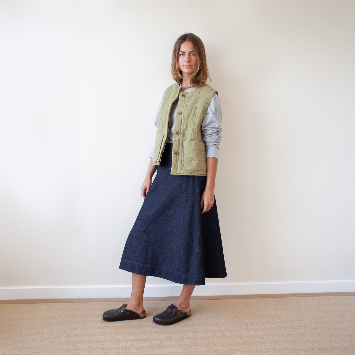 A woman stands in a minimalist room with a white wall and light wood floor, wearing a light olive green quilted vest over a light gray long-sleeve top and a dark navy midi skirt, paired with dark brown mule shoes.