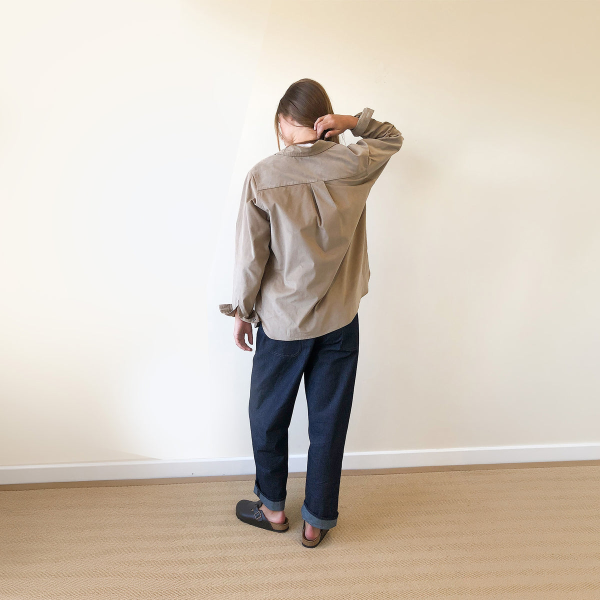 A person standing with their back to the camera in a minimalist room with a beige wall and light wood floor, wearing a light beige button-up shirt, dark navy pants, and dark shoes.