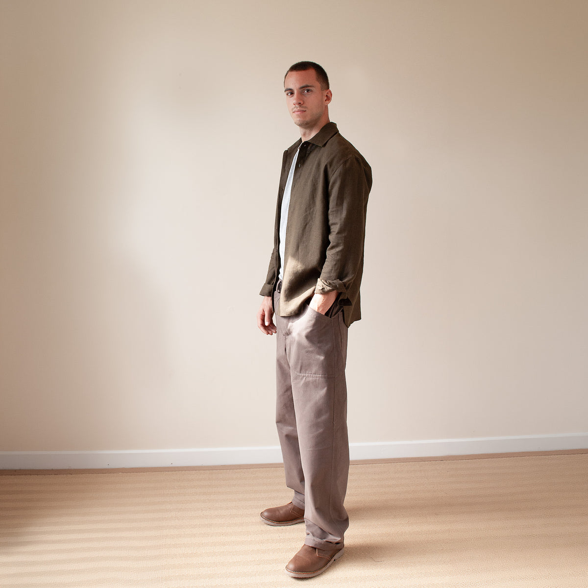 A man wearing a dark olive green overshirt, white t-shirt, taupe trousers, and brown shoes, standing in profile against a plain beige wall and light wood floor.
