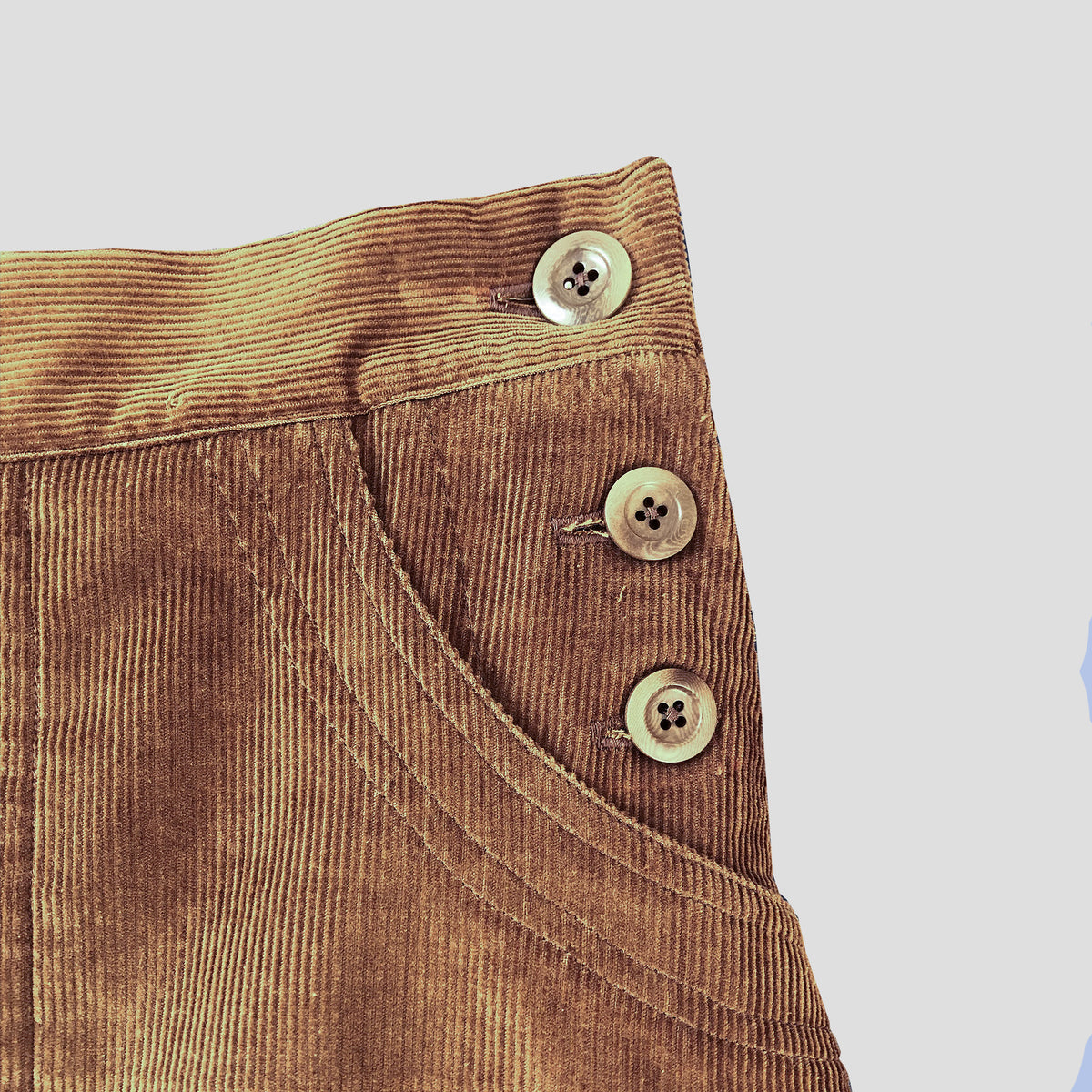 Close-up of a brown corduroy skirt, showing the fabric texture, a pocket seam, and three decorative buttons on a light gray background.