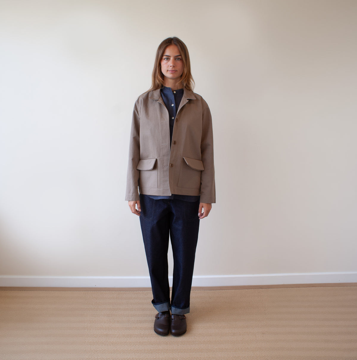 A woman stands against a plain beige wall on a light wood floor, wearing a taupe utility jacket over a navy shirt, dark blue jeans, and dark brown shoes.