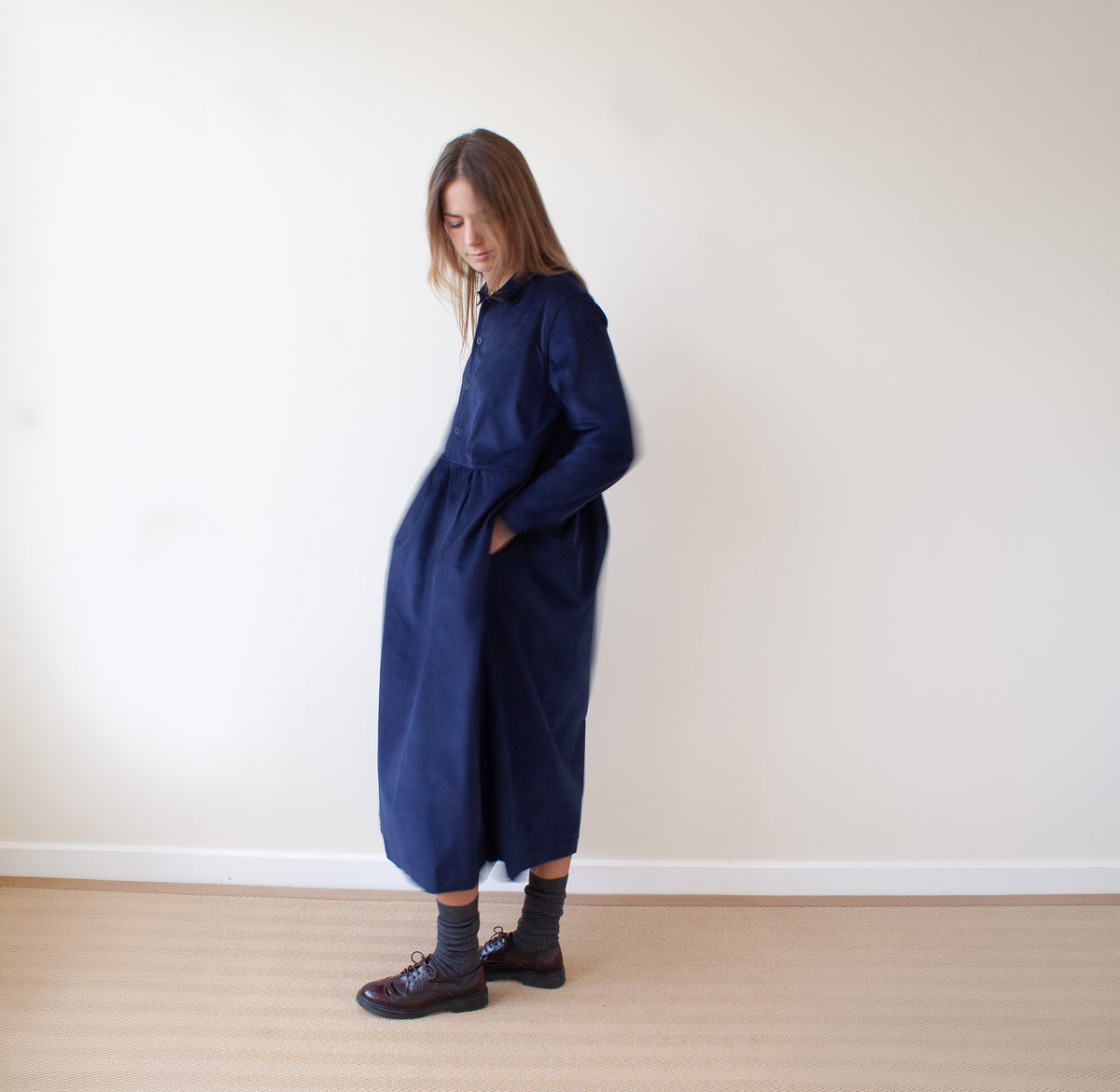 A woman wearing a long navy blue dress with side pockets, styled with dark gray socks and brown lace-up shoes, standing in a minimalist room with a cream wall and light wood floor.