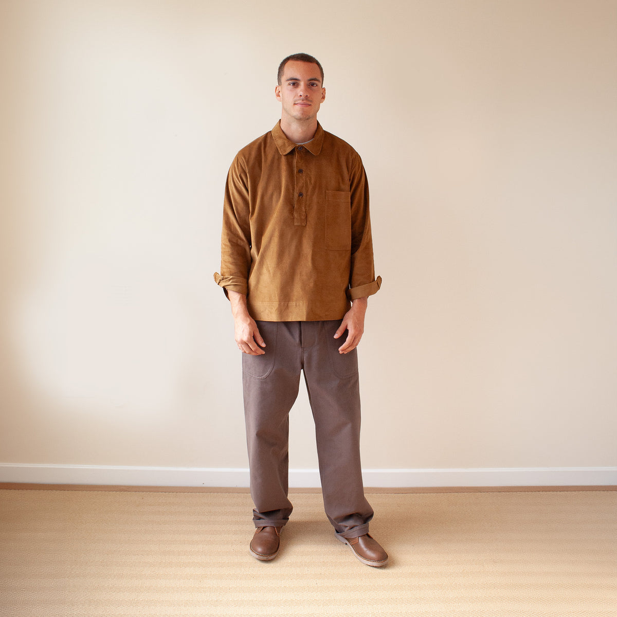A man stands in a minimalist room with a beige wall and light wood floor, wearing a brown corduroy long-sleeve button-up shirt, brown trousers, and brown leather shoes.