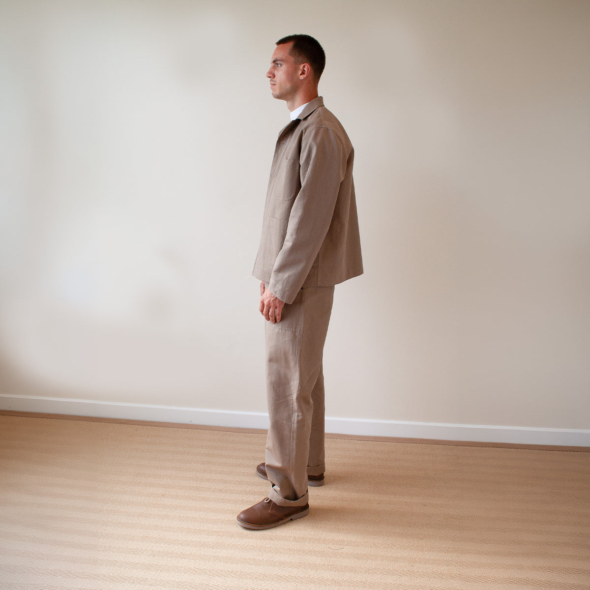 A man in a side profile wearing a light brown linen suit, consisting of a jacket and trousers, paired with brown leather shoes, standing in a minimalist room with a beige wall and light wood floor.