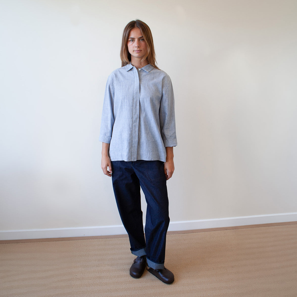 A woman stands in a minimalist room with a beige wall and light wood floor, wearing a light blue linen button-up shirt and dark navy wide-leg jeans.