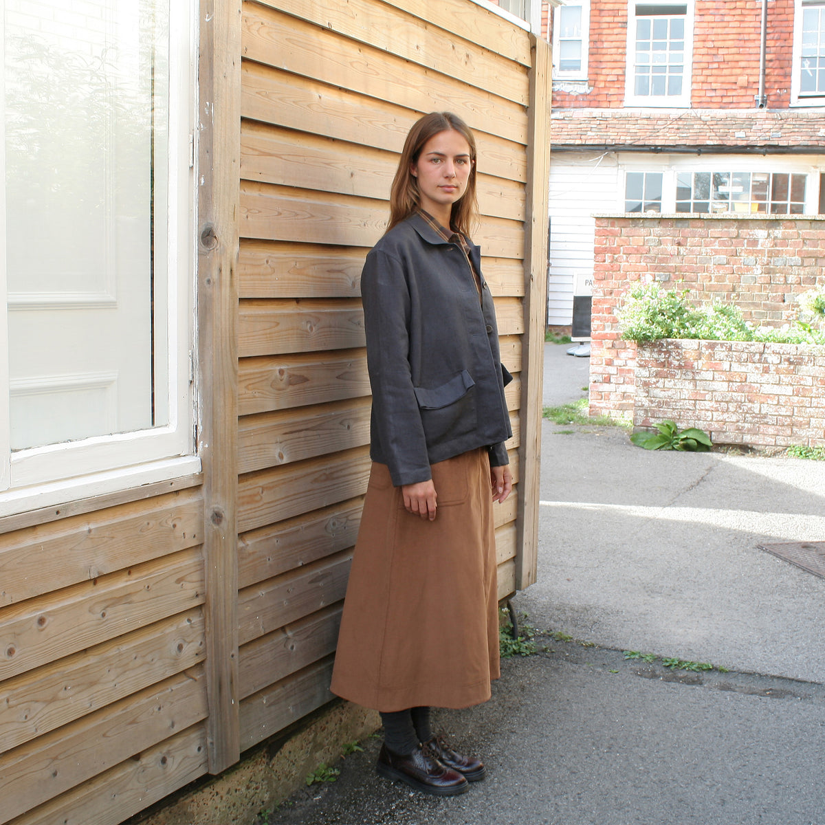 A woman stands outdoors against a wooden building wall, wearing a dark charcoal utility jacket and a brown midi skirt.
