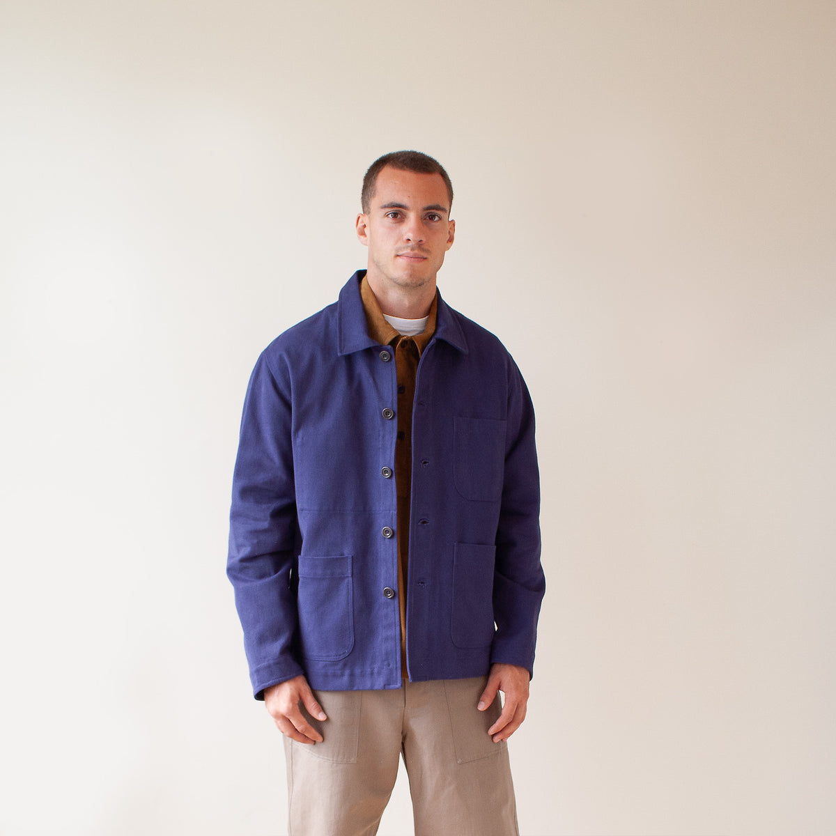 A man wearing a navy blue utility jacket over a brown shirt and beige pants, standing against a plain beige background.