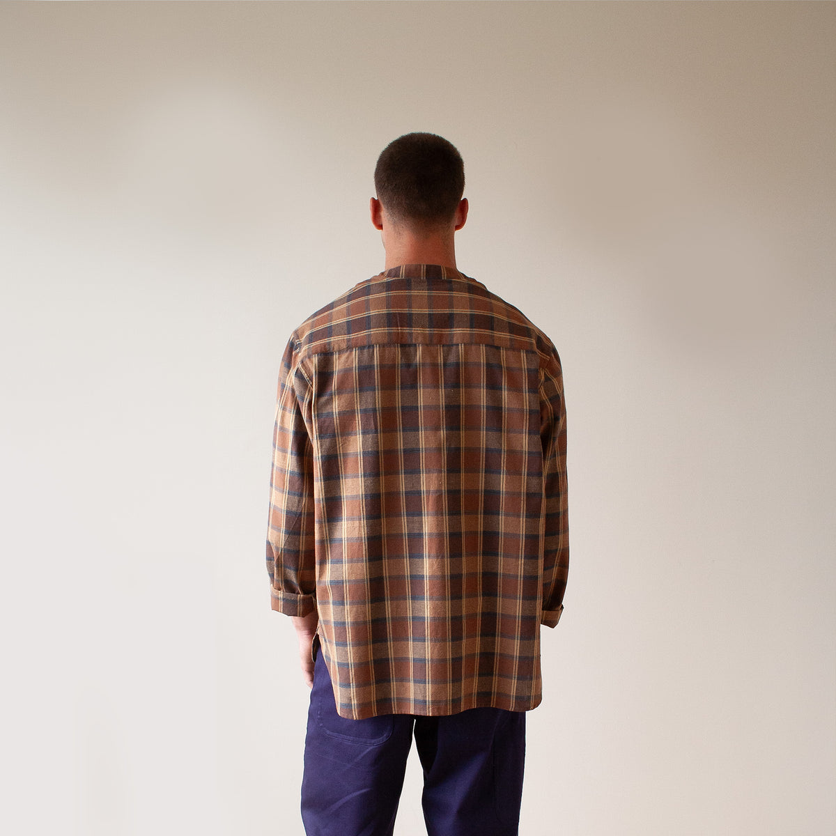 A man viewed from behind wearing a brown and beige plaid button-down shirt and dark blue pants, standing against a plain light beige wall.