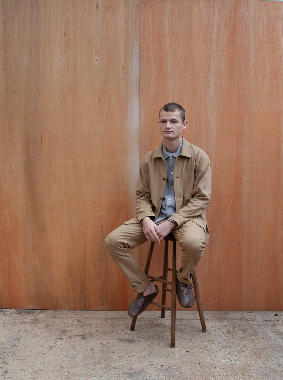 a young man standing against a wooden wall wearing a traditional chore jacket made in a sustainable tan brushed cotton canvas