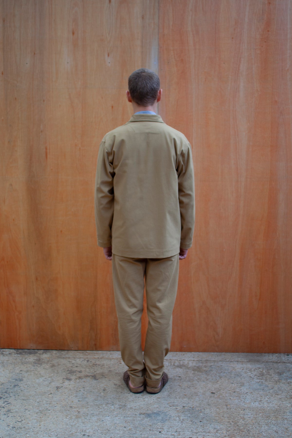 a young man standing against a wooden wall wearing a traditional chore jacket made in a sustainable tan brushed cotton canvas