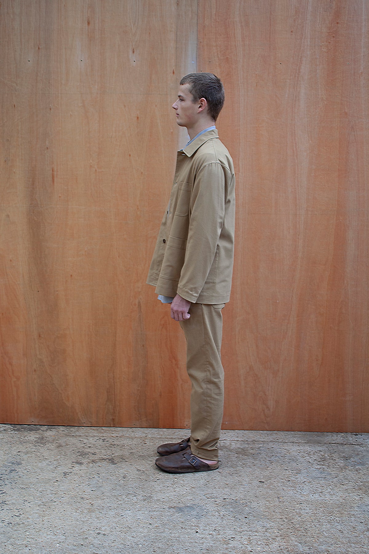 a young man standing against a wooden wall wearing a traditional chore jacket made in a sustainable tan brushed cotton canvas
