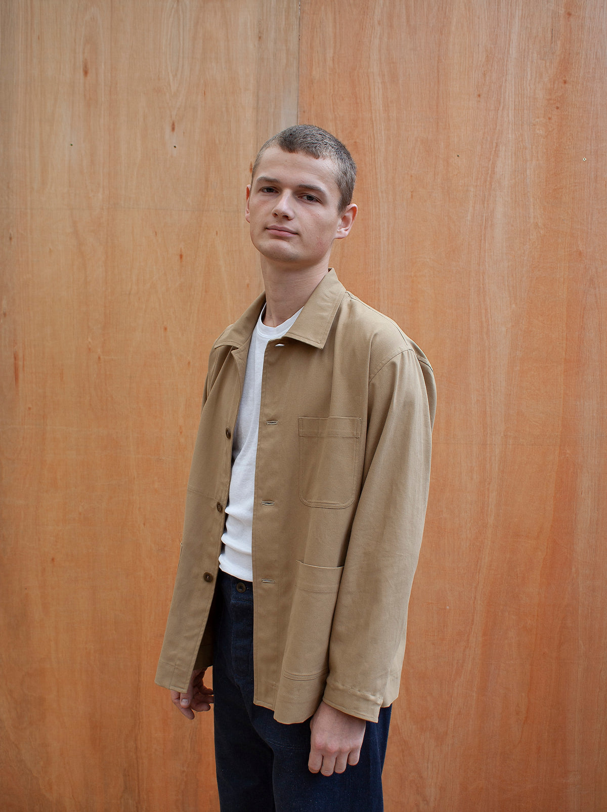 a young man standing against a wooden wall wearing a traditional chore jacket made in a sustainable tan brushed cotton canvas
