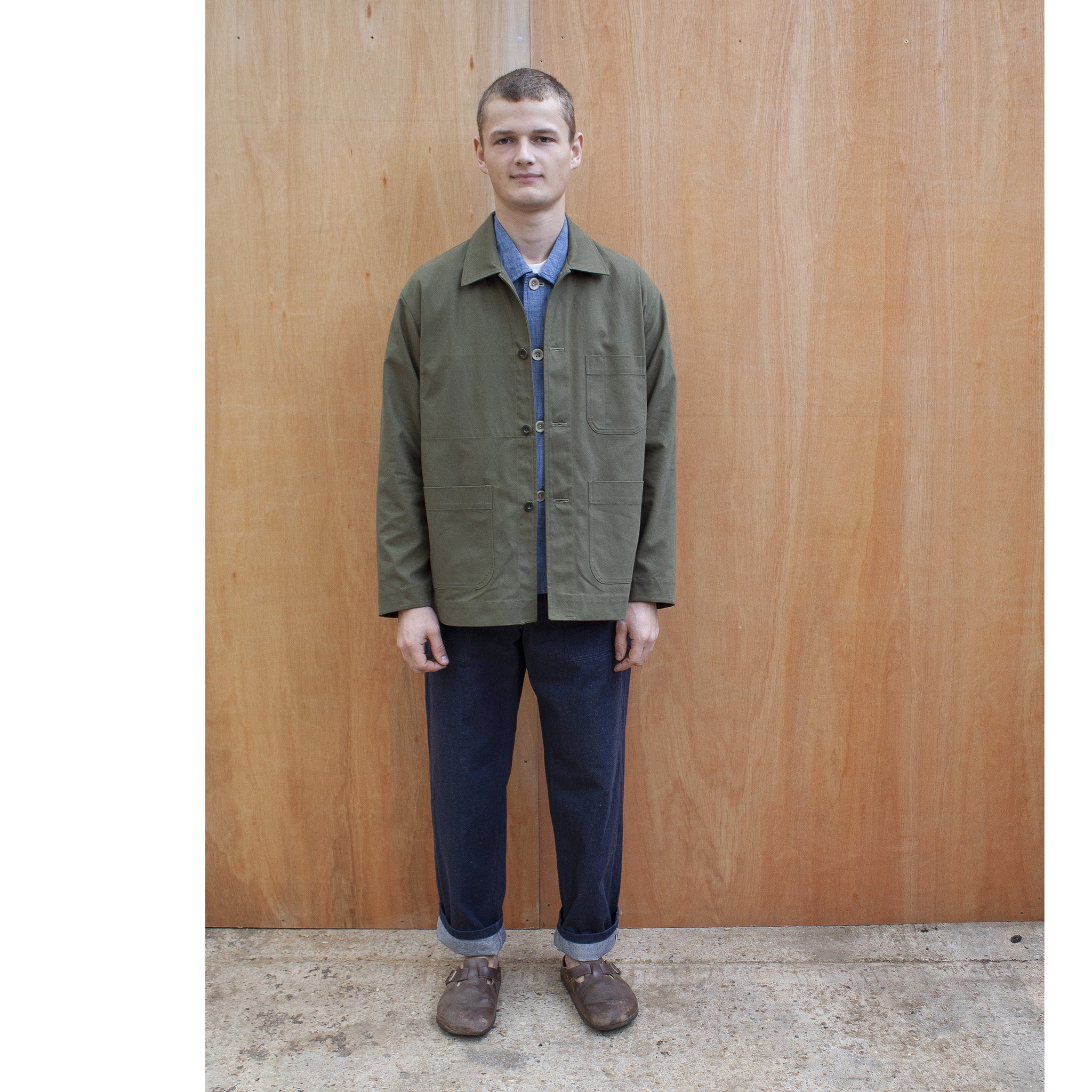 a young man standing against a wooden wall wearing a traditional chore jacket made in a sage green brushed cotton canvas