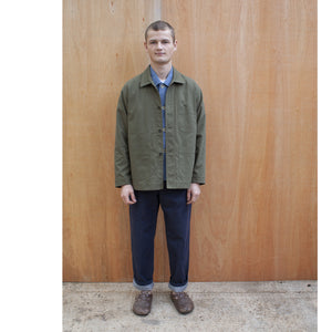 a young man standing against a wooden wall wearing a traditional chore jacket made in a sage green brushed cotton canvas