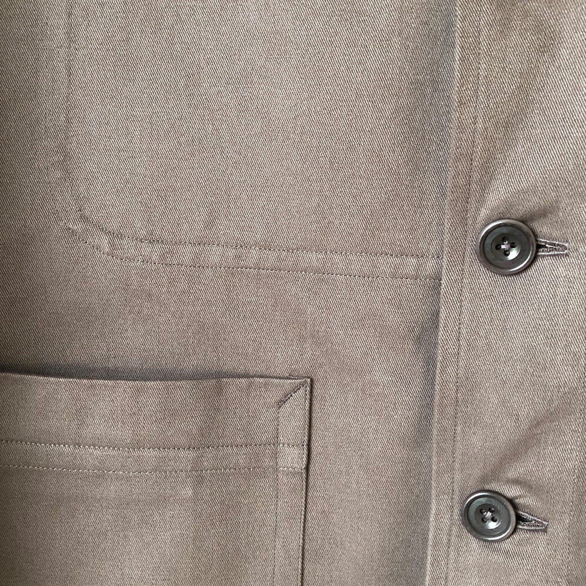 Close-up of a light taupe-beige textured fabric garment showing a pocket, button closure, and fine woven texture.