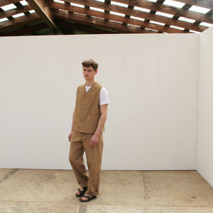 a young man stands in a white space wearing a tan waistcoat and matching chore trousers, with a short sleeved white t-shirt