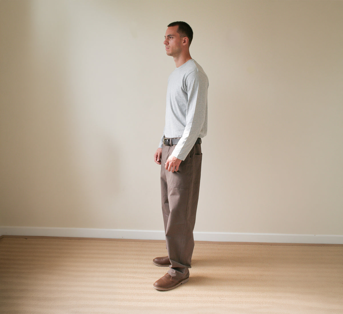 A man standing in profile wearing a light gray long-sleeve shirt, brown trousers, and brown shoes in a minimalist room with beige walls and light wood flooring.