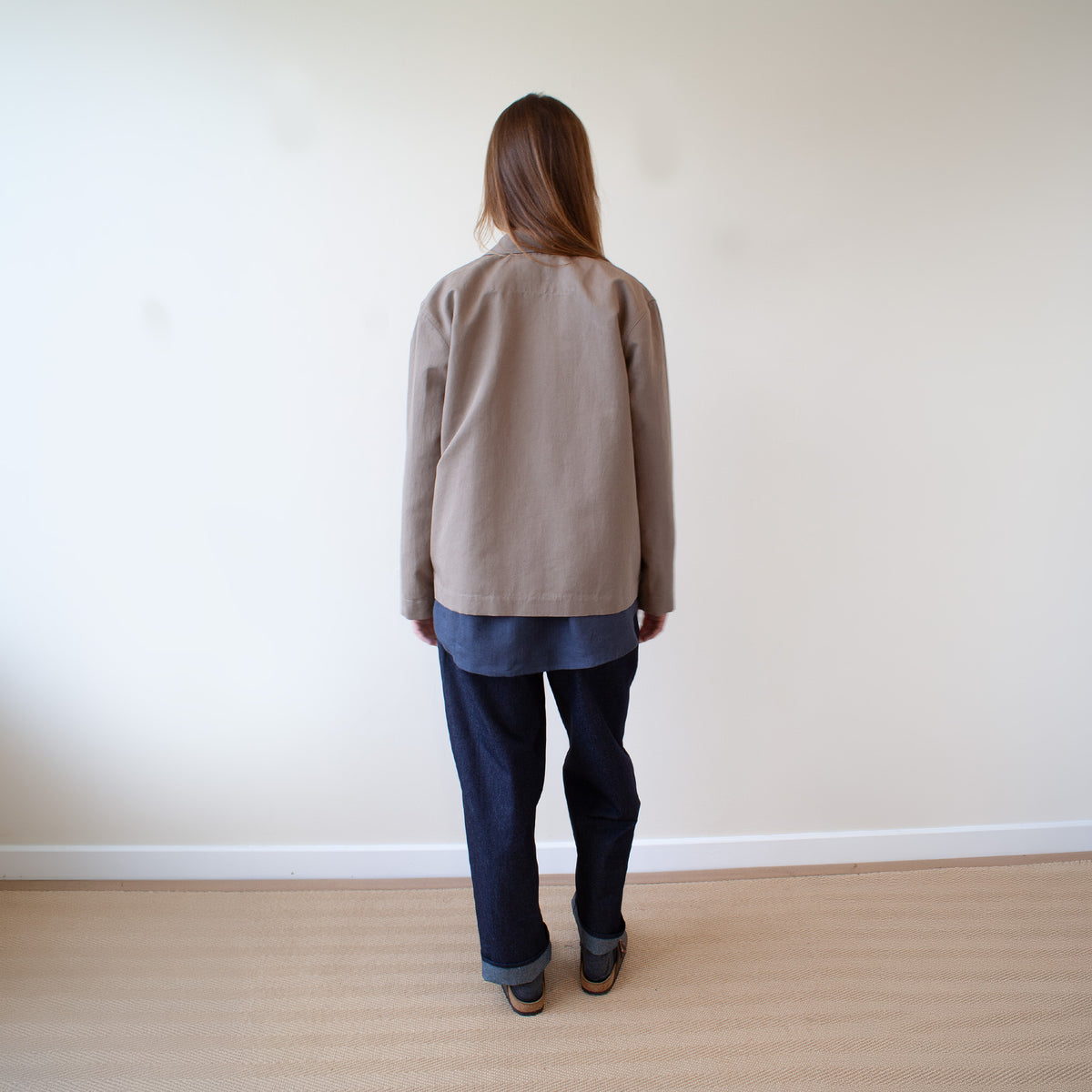 A woman is shown from behind wearing a beige long-sleeve linen jacket over a blue shirt and dark blue jeans, standing on a light wood floor against a plain off-white wall.