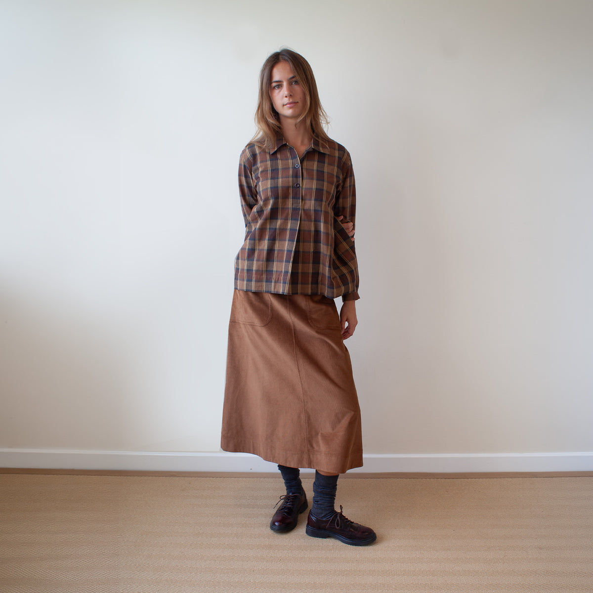 A woman stands in a minimalist room with a beige wall and light wood floor, wearing a brown and orange plaid button-down shirt and a brown midi skirt.