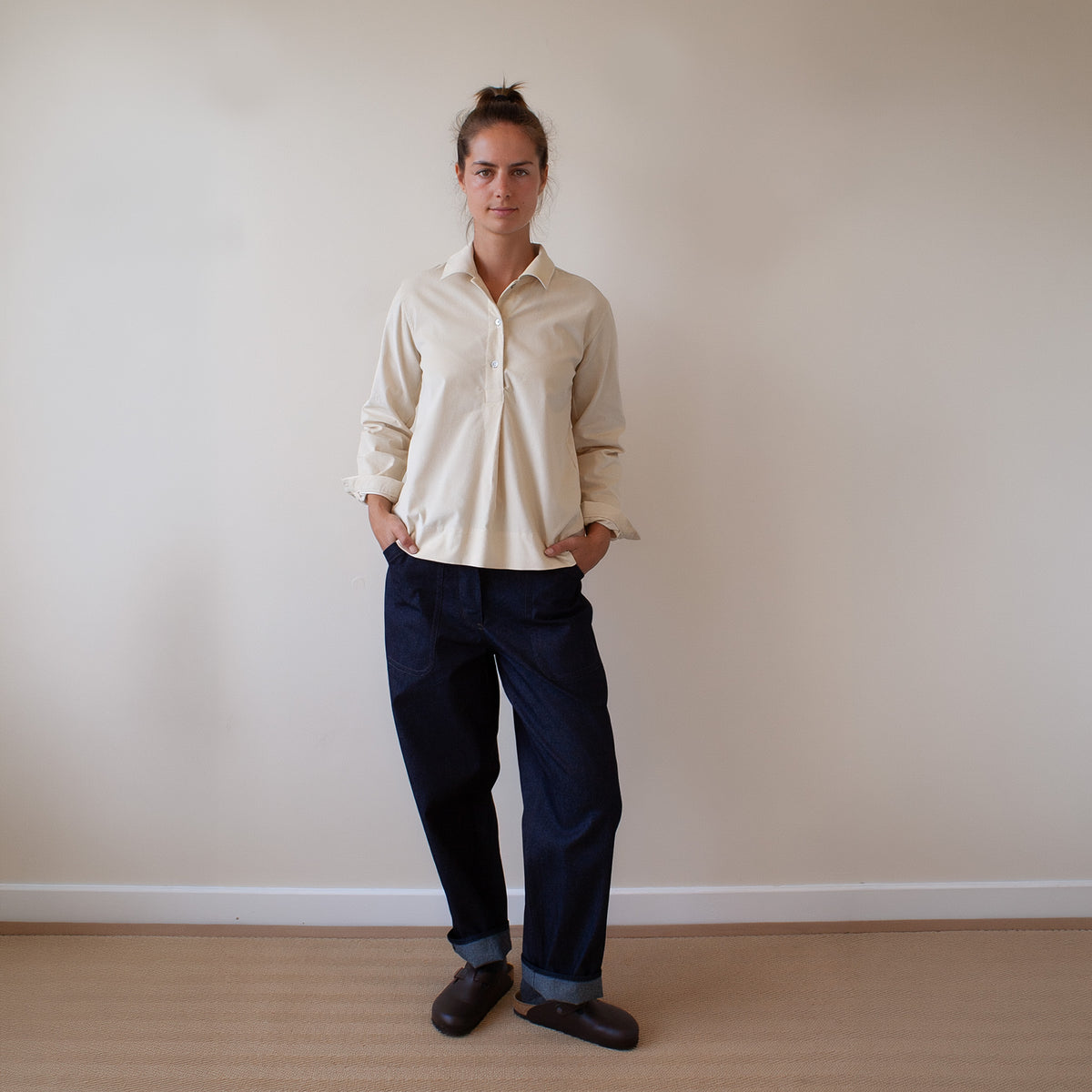 A woman stands against a plain beige wall wearing a light cream button-up shirt, dark navy trousers, and dark brown shoes.