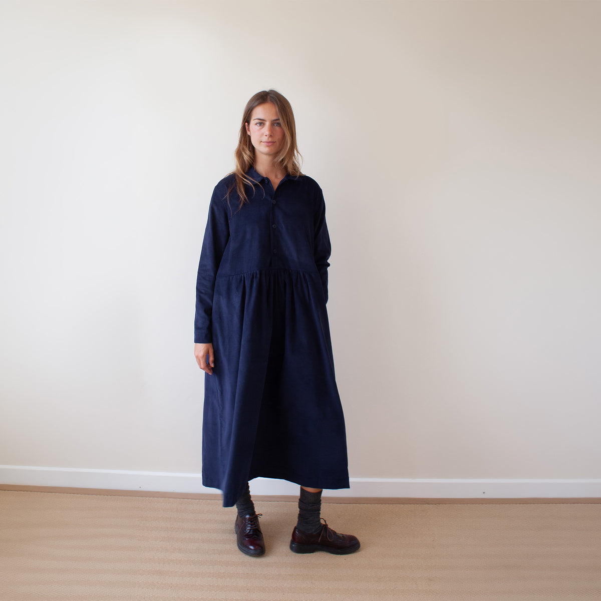A woman stands in a minimalist room with a beige wall and light wood floor, wearing a long navy blue corduroy dress with a button-up front and long sleeves.