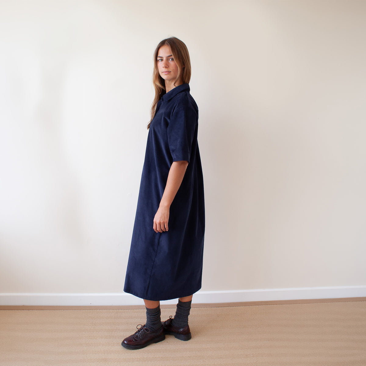 A woman wearing a dark navy blue corduroy midi dress with short sleeves, standing in profile against a plain beige wall and light wood floor.