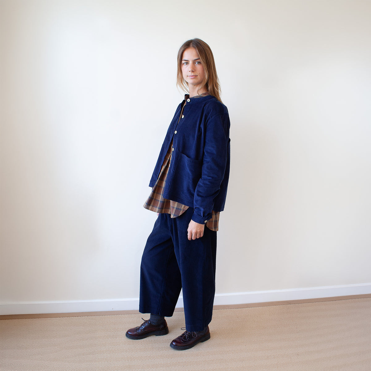 A woman stands in profile wearing a dark navy corduroy jacket with a brown and beige plaid shirt underneath and matching navy corduroy trousers, set against a plain off-white wall and light wood floor.
