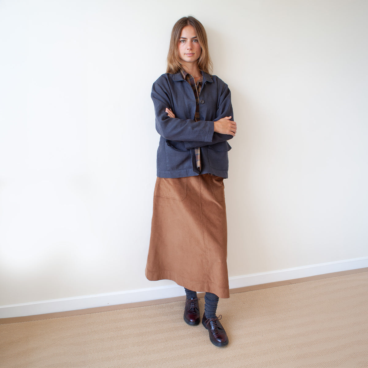 A woman stands with arms crossed, wearing a dark blue button-up shirt, a brown midi skirt, dark socks, and dark leather shoes, posing against a plain off-white wall on a light wood floor.