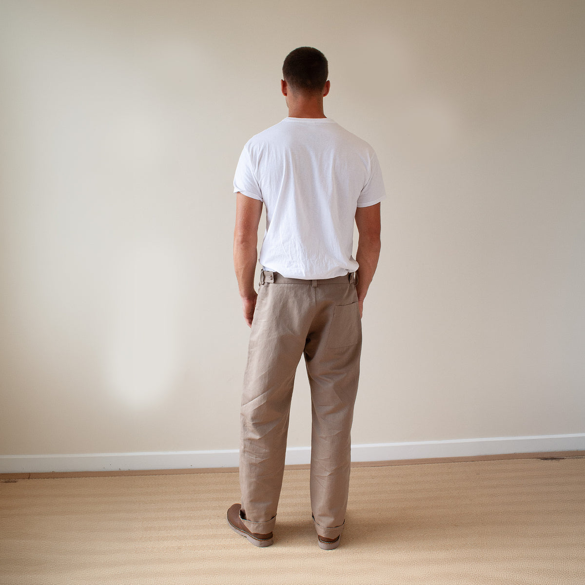 A man viewed from behind wearing a white t-shirt and beige trousers, standing on a light wood floor against a plain beige wall.