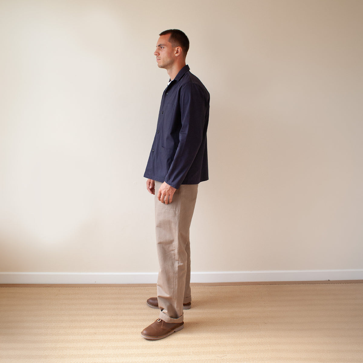 A man standing in profile wearing a dark navy long-sleeve button-up shirt, beige trousers, and brown slip-on shoes, set against a plain beige wall and light wood floor.