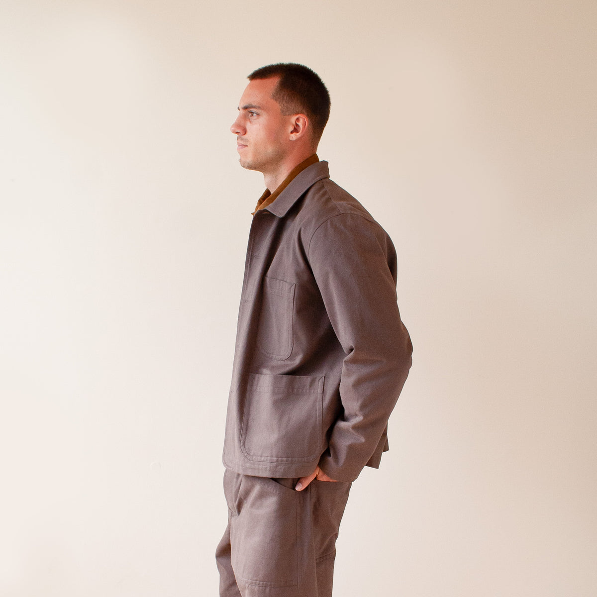 A man in profile wearing a dark brown utility jacket and matching trousers against a plain beige background.