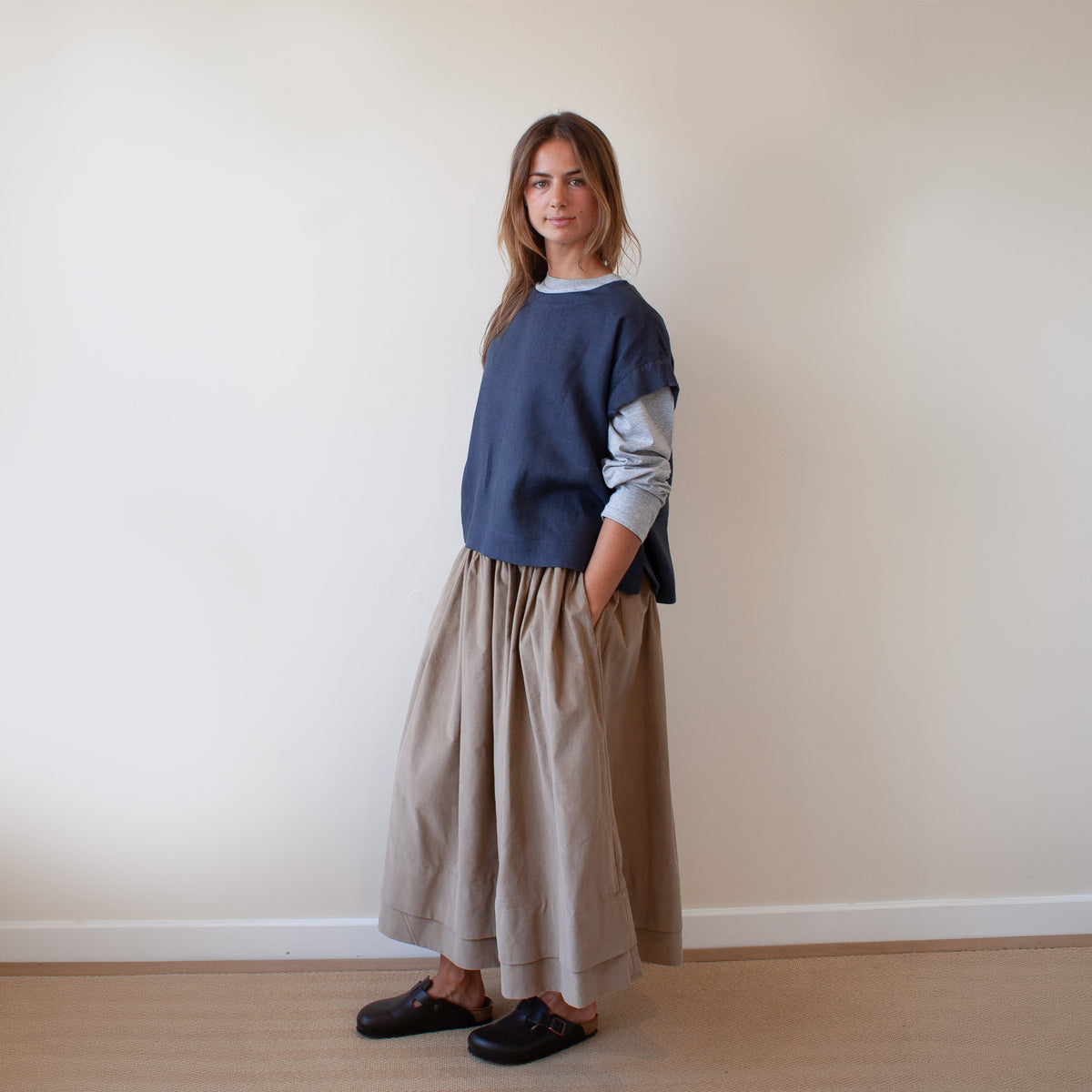 A woman stands in a minimalist room wearing a navy blue crewneck sweater layered over a light gray long-sleeve shirt, paired with a beige linen skirt and dark loafers.