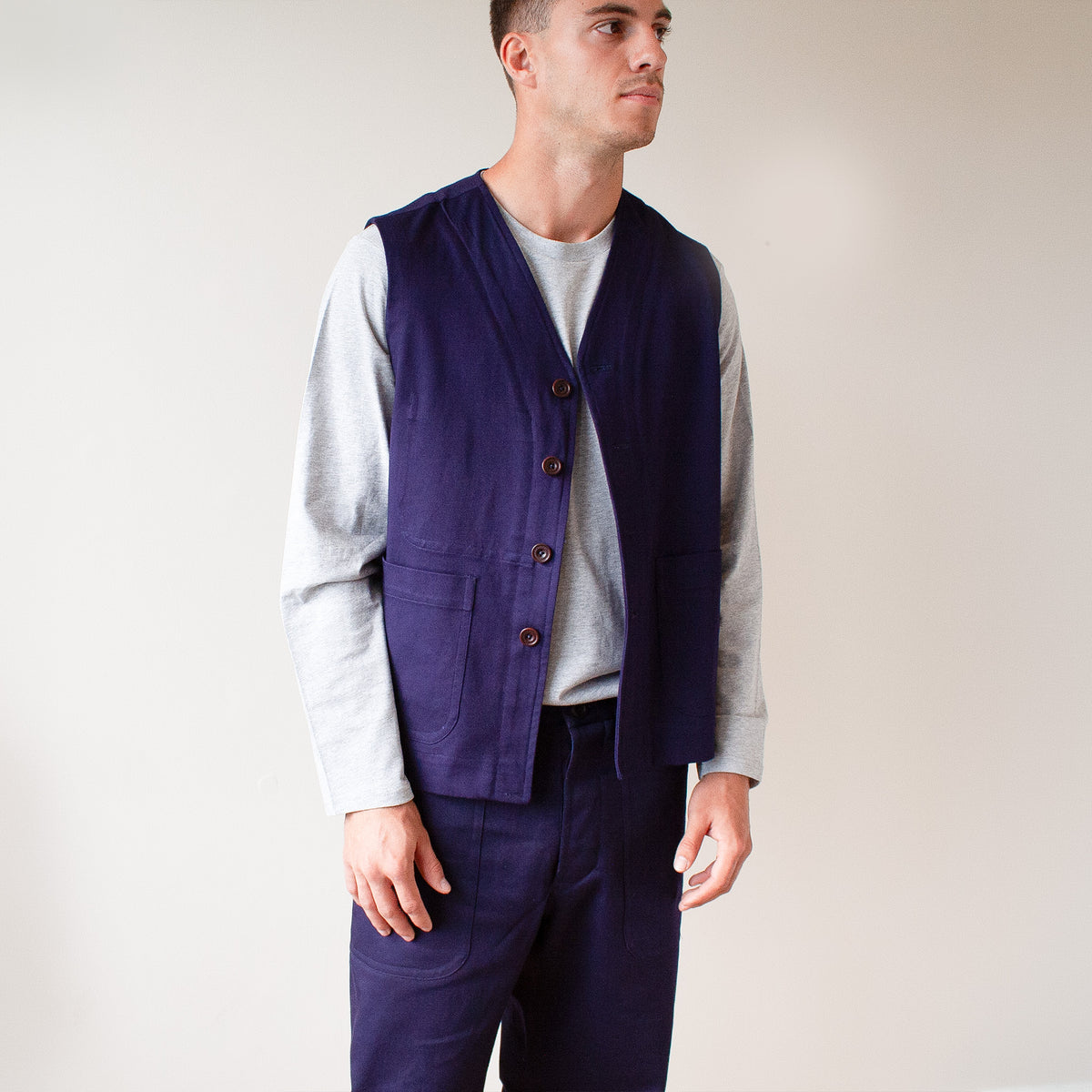 A man wearing a dark navy blue linen vest over a light gray long-sleeve shirt and matching navy trousers, standing against a plain off-white background.
