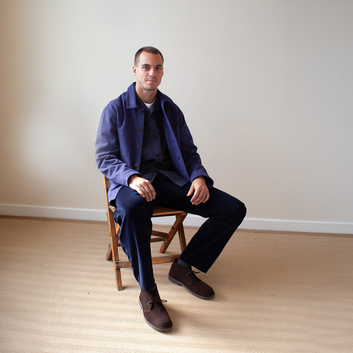 A man sits on a wooden folding chair in a minimalist room with a beige wall and light wood floor, wearing a navy blue overshirt over a dark shirt and trousers with brown suede shoes.