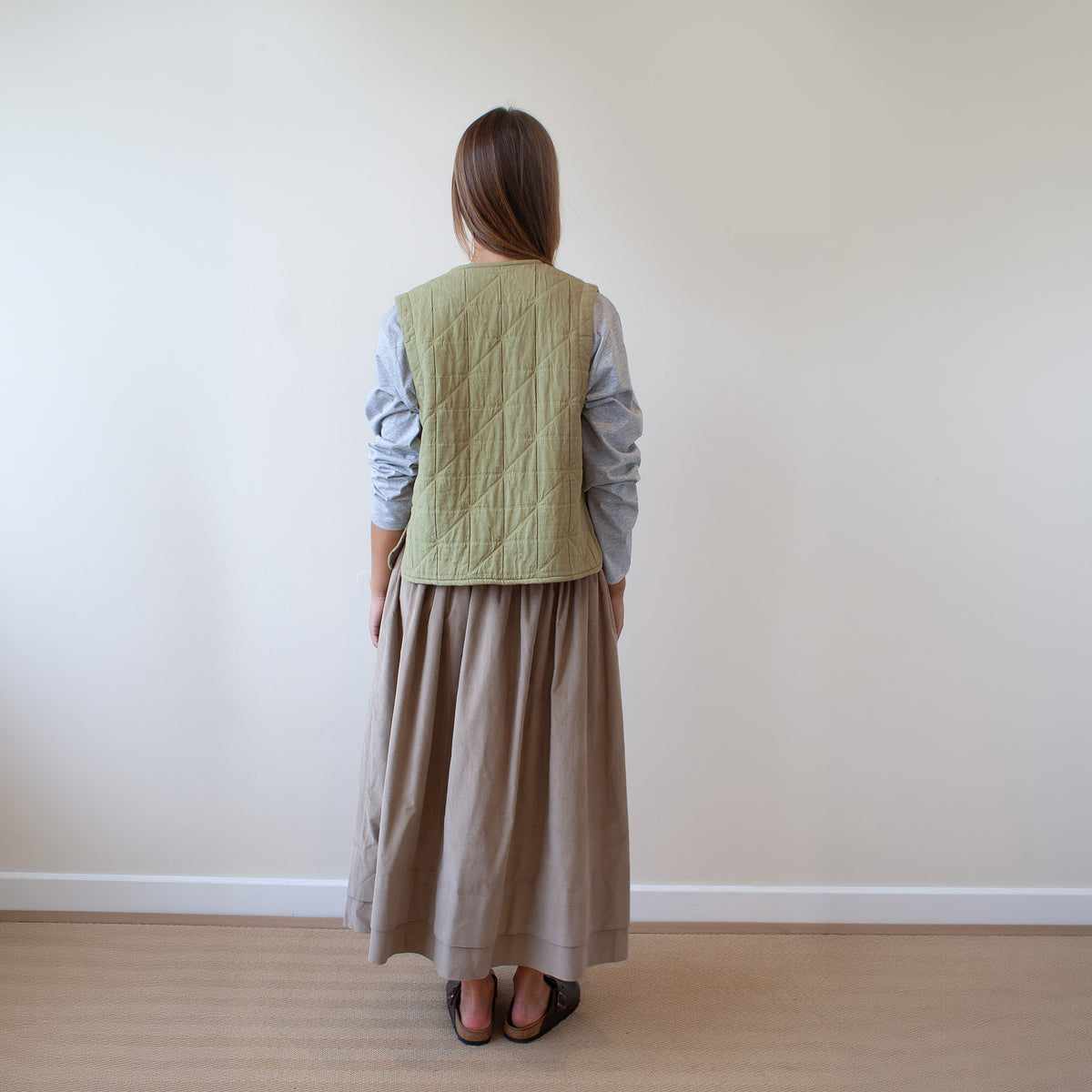 A woman viewed from behind wearing a light sage green quilted vest over a light blue long-sleeve top and a long taupe skirt, standing on a light wood floor against a plain beige wall.