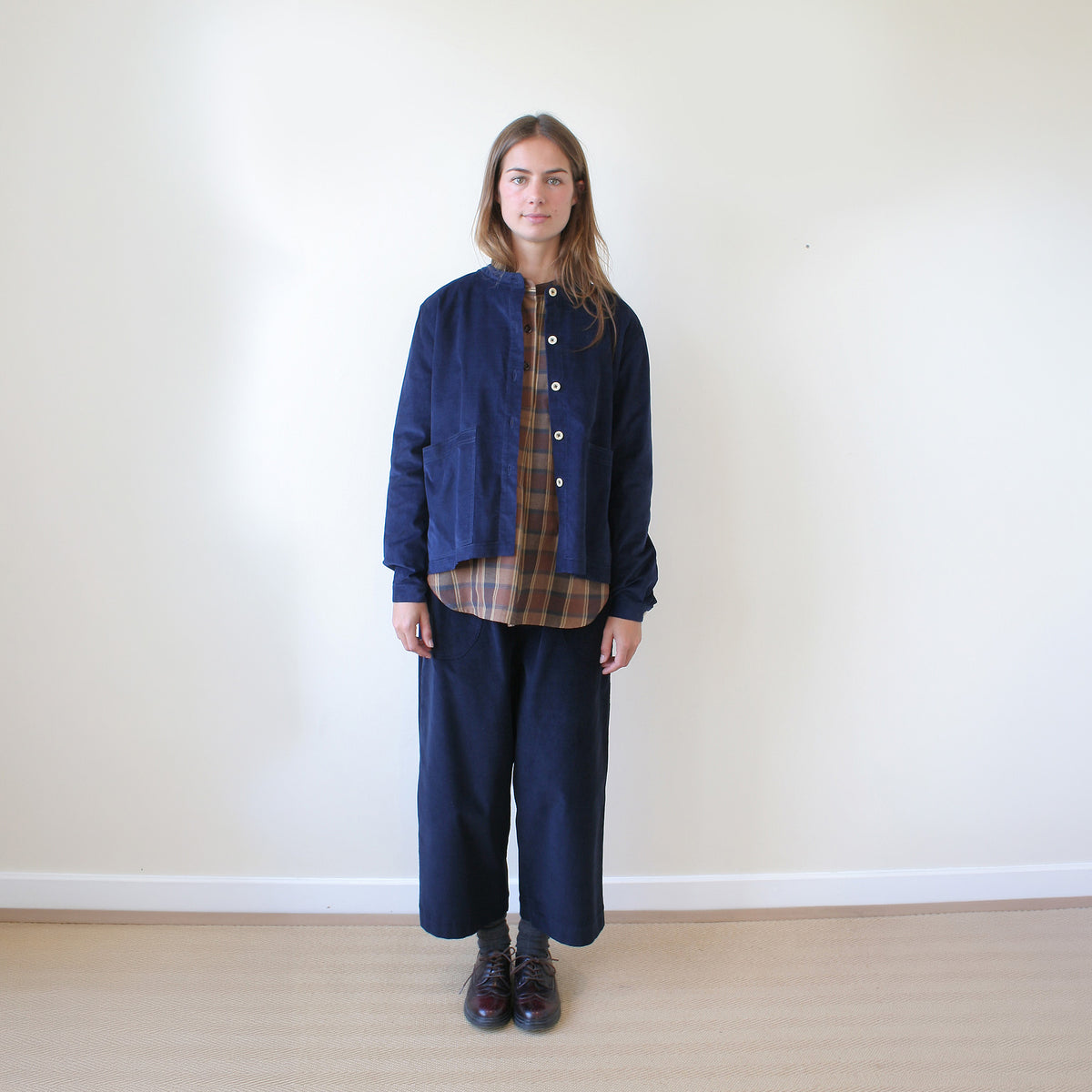 A woman stands against a plain off-white wall wearing a navy blue corduroy jacket over a brown and beige plaid shirt, paired with matching navy corduroy trousers and dark brown shoes.