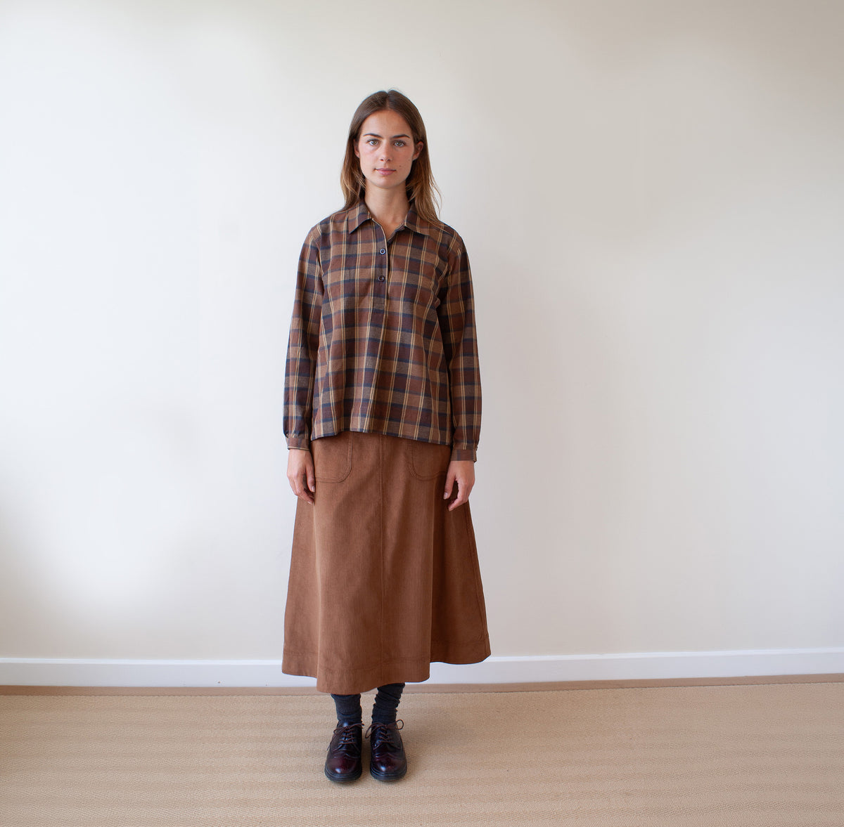 A woman stands against a plain off-white wall wearing a brown and beige plaid button-up shirt and a brown midi skirt, paired with dark socks and black shoes.
