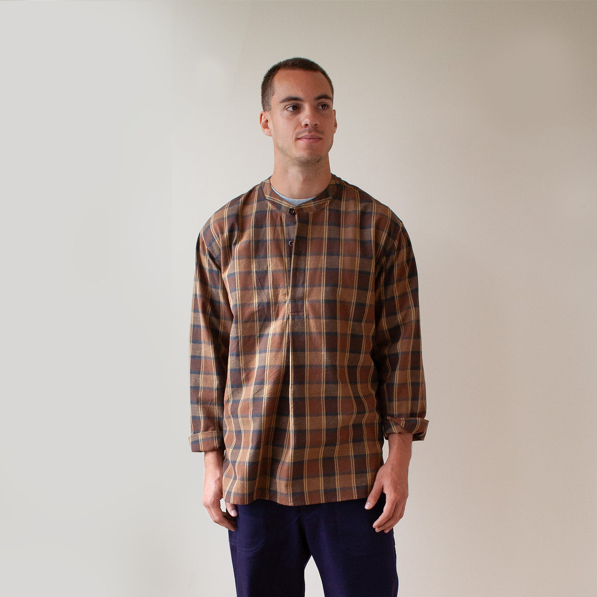 A man wearing a brown and beige plaid long-sleeve shirt with a band collar, standing against a plain light beige wall.