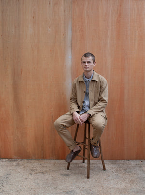 a young man standing against a wooden wall wearing a traditional chore jacket made in a sustainable tan  brushed cotton canvas