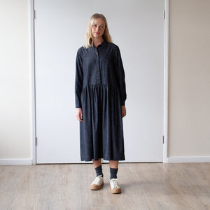 young blonde woman looking forward stands against a cream wall wearing a simple grey denim dress with a gathered waist and long sleeves