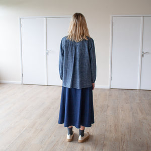 young blonde woman stands in a sunlit room, her back is towards us and you can see a deep back pleat detail on the shirt