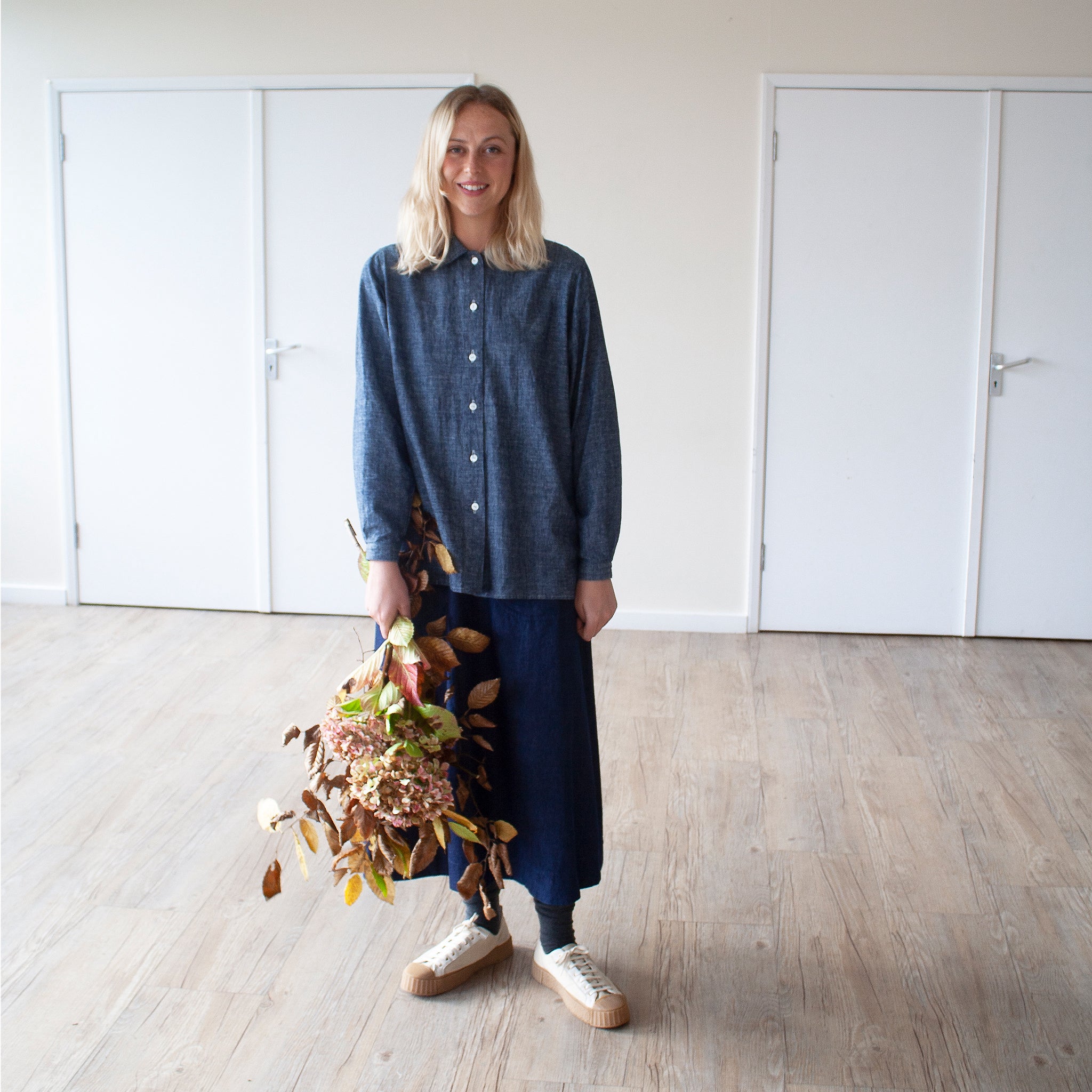 smiling blonde young woman standing in a sunlit room wearing a blue cotton hemp shirt, she is holding s large bunch of autumnal leaves and flowers