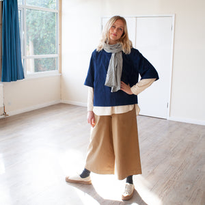 young blond woman wearing a pair of elasticated tan 3/4 trousers with a blue top and scarf standing in a sunlit room