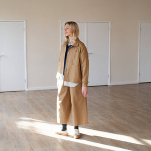 young blond woman standing in a sy=unlit room, wearing a tan cotton casual jacket and trousers