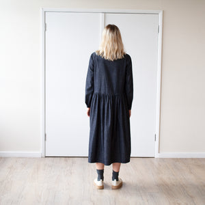 young blonde woman looking backwards stands against a cream wall wearing a simple grey denim dress with a gathered waist and long sleeves