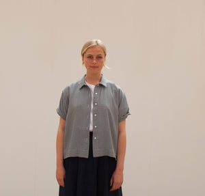 A young blond woman facing the camera wearing a ging=ham short sleeved shirt and a navy linen skirt