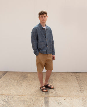 A young man stands in a white space wearing a light blue denim chore jacket and a pair of tan coloured cotton shorts
